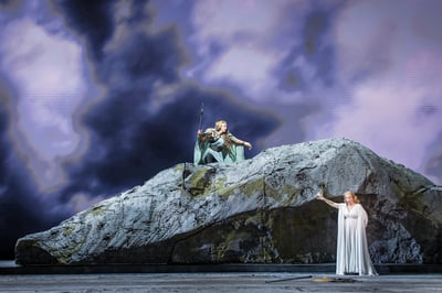 A figure with a spear lunges from the summit of the rock toward a woman in white below, under a full-stage wall of storm-cloud projections.
