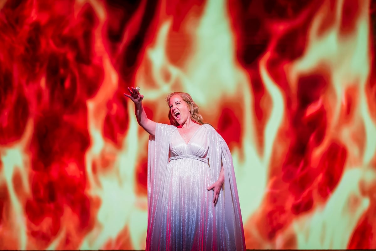 Brünnhilde in a silver-white gown raises the ring against a full-stage wall of projected flame in the Immolation Scene.