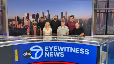 Group photo of the lighting + install crew at the KABC ABC7 Eyewitness News anchor desk after the studio install, LA skyline backdrop visible through the upstage windows