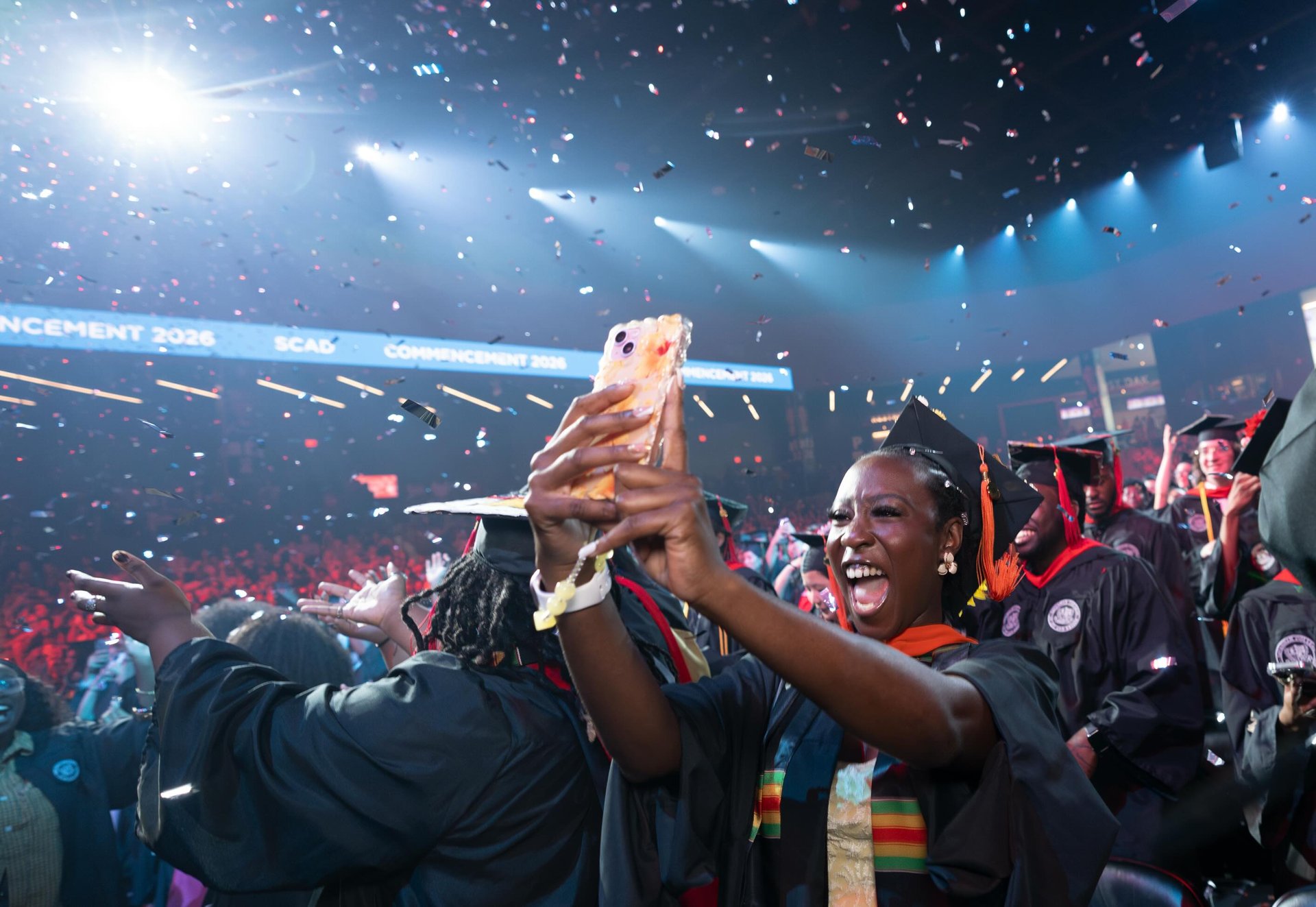 A SCAD graduate in cap and gown beams while raising a phone to film amid falling confetti, the blue-lit arena bowl and a glowing 'Commencement 2026' LED ribbon behind a crowd of fellow graduates.