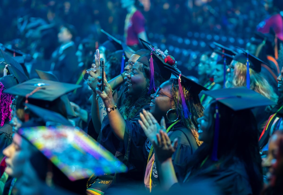 Rows of SCAD graduates in caps and gowns under a deep blue wash, several lifting phones to capture the ceremony, tassels marked 2026.