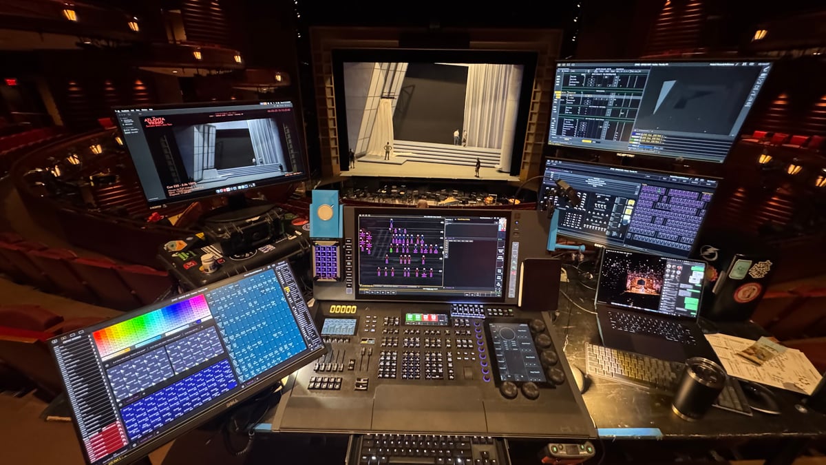 Wide view of the Eos programming desk from the orchestra during Semele tech, full setup with console, dual XKeys, and multiple monitors, with the white-curtain-and-pillar Wedding Hall set visible on stage behind