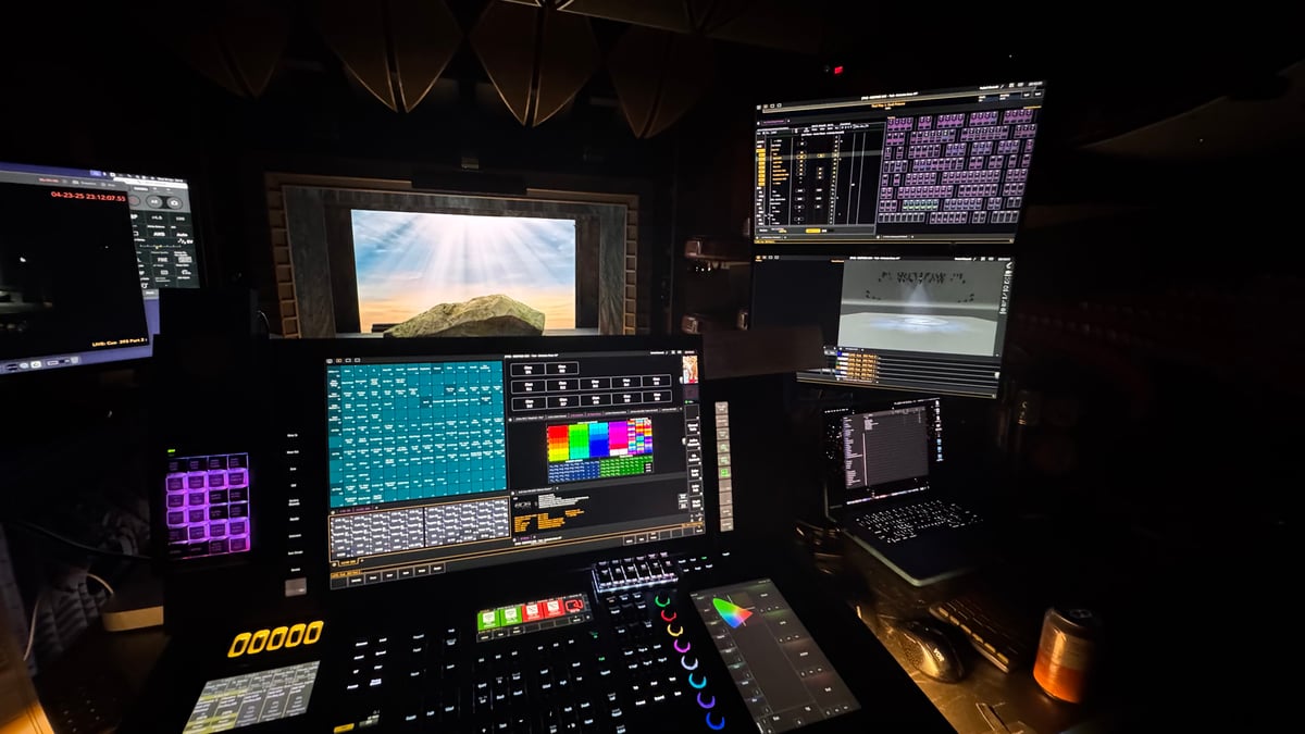 Full programming desk during Siegfried tech, Eos console with dual XKeys panels and multiple monitors showing magic sheet, color palette, and cue list, with the Act III sunrise mountain projection visible on the stage screen behind