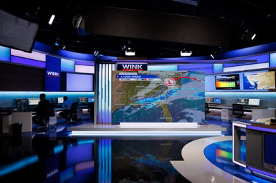 Wide view of WINK News' new weather set at the Gateway broadcast center, anchor desk with full-wall weather display, meteorologist consoles to the side, deep blue practical accents reflecting off the polished floor