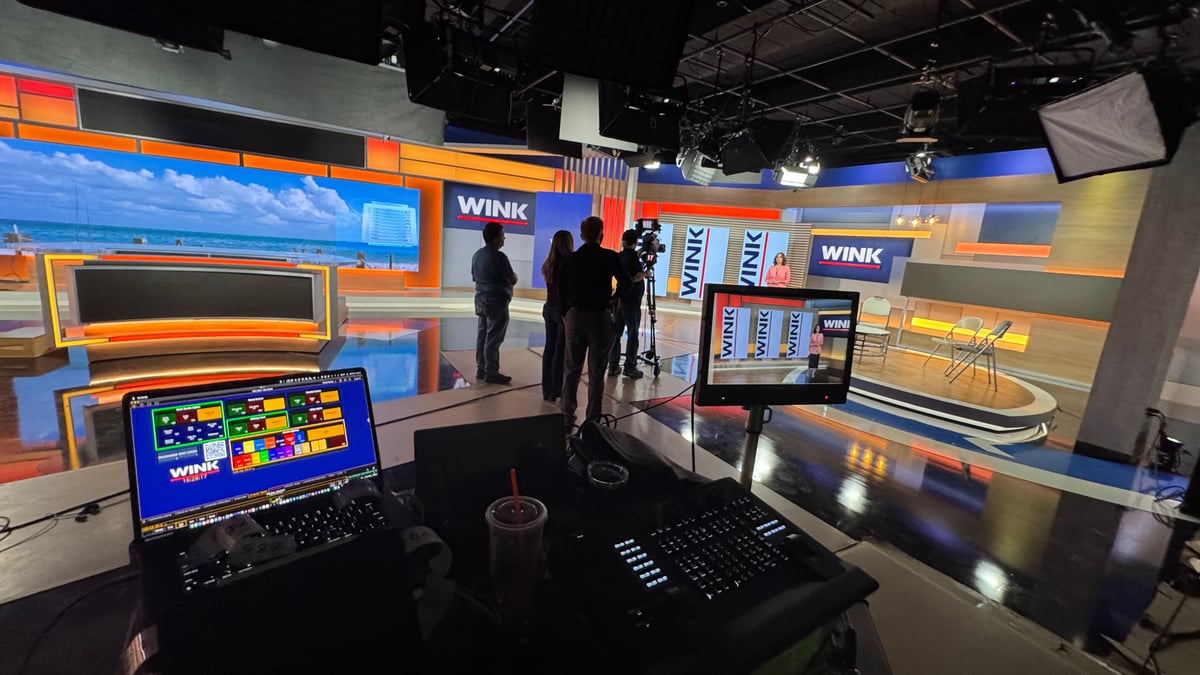 Behind-the-scenes from the WINK install, Mike's programming laptop in the foreground, the broadcast set + crew + anchor visible past the camera, daylight from the weather-wall beach scene flooding the studio