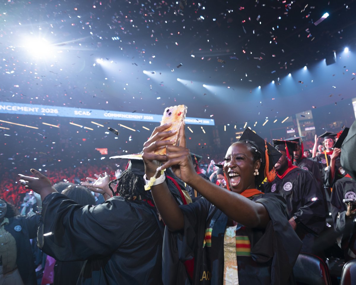 A SCAD graduate cheers with her phone raised as confetti rains down through a bright spotlight beam and a hazy magenta wash at the commencement finale.