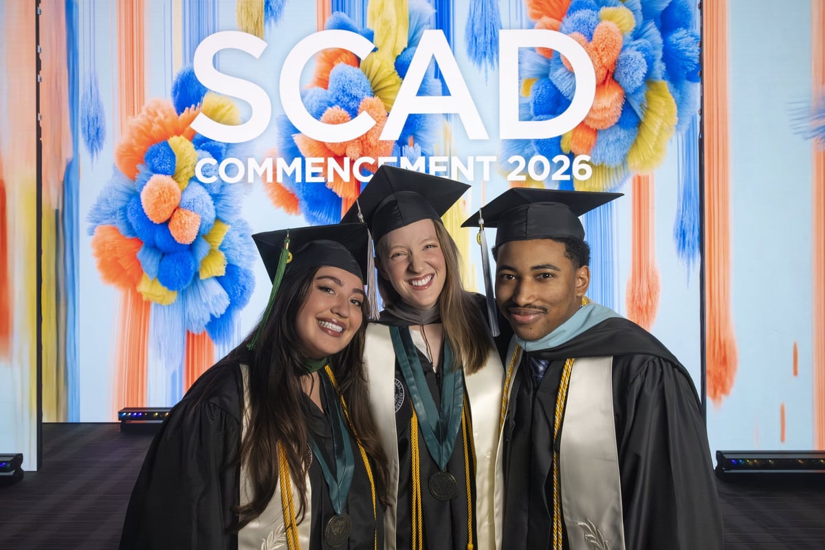 Three SCAD graduates in caps and gowns pose together, smiling, in front of a vivid blue and orange painted backdrop reading 'SCAD Commencement 2026.'