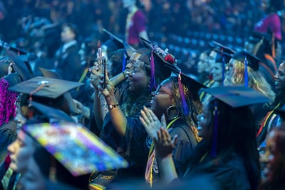 Graduating students fill the arena under a deep blue wash, one lifting her phone to film the ceremony from her seat.