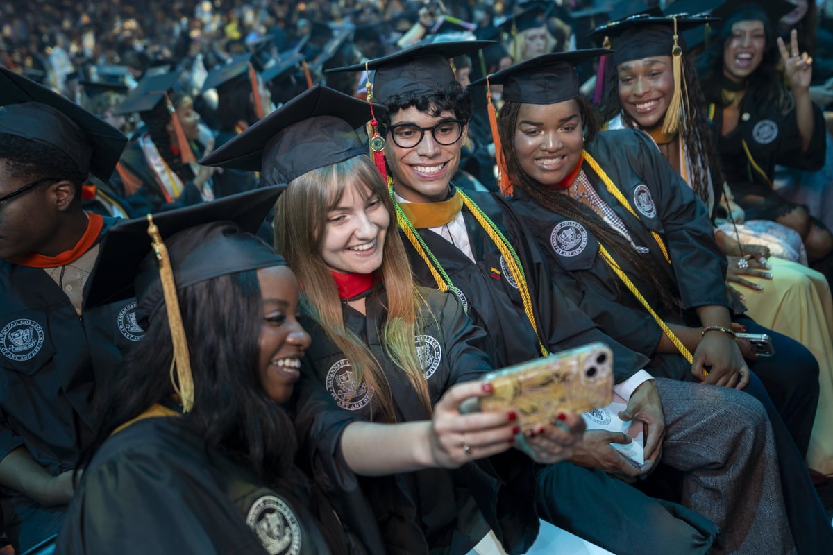 A row of seated graduates leans in for a group selfie on a gold phone, warmly lit against the packed arena behind them.