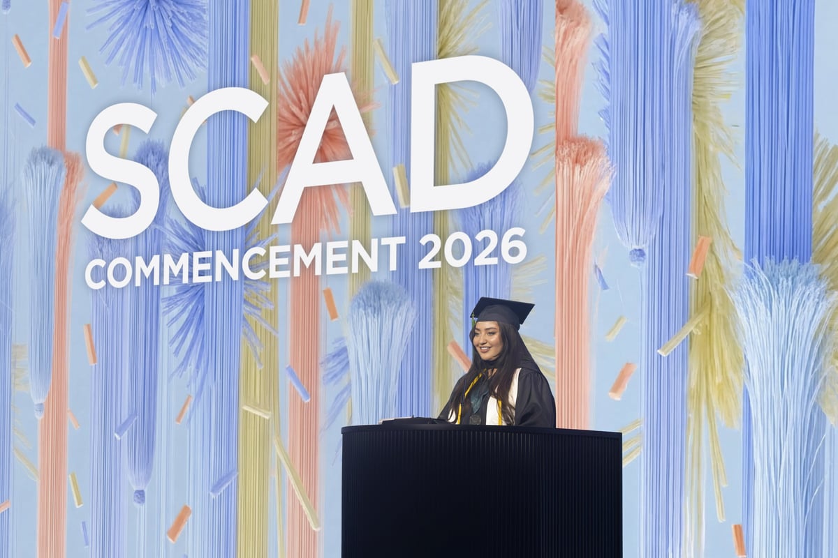 A graduate speaks from a black podium in front of a soft pastel backdrop reading 'SCAD Commencement 2026,' lit in cool blue and coral tones.