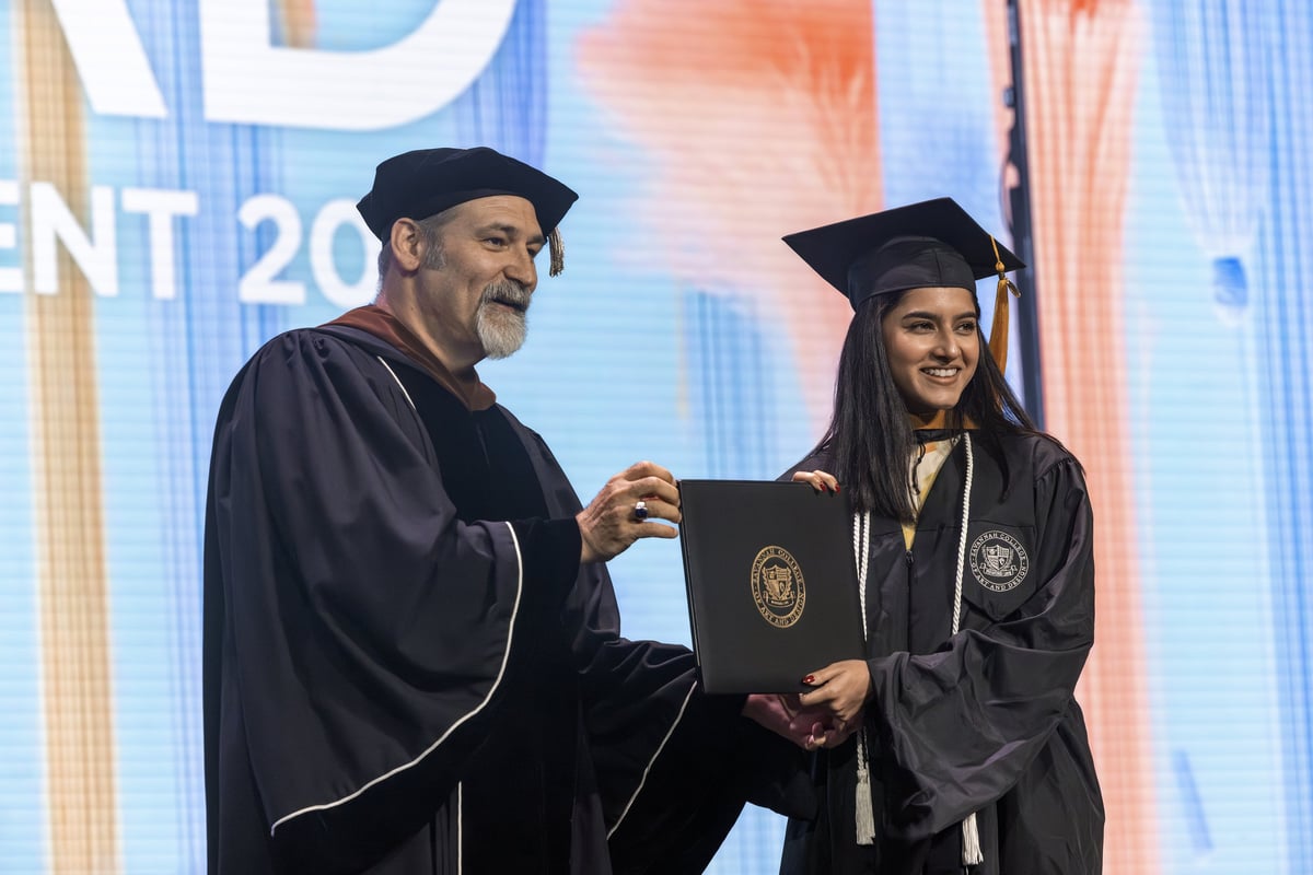 A graduate shakes hands with a faculty member in academic regalia as she receives her diploma on stage, both smiling in front of the pastel 'SCAD Commencement 2026' backdrop.