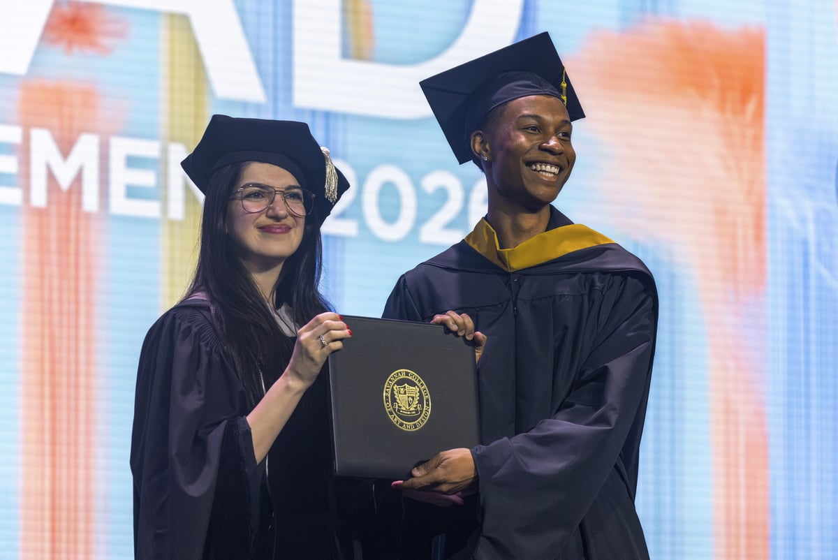 A master's graduate in a gold-trimmed hood poses with a presenter, both holding his SCAD diploma cover and smiling in front of the 'SCAD Commencement 2026' backdrop.