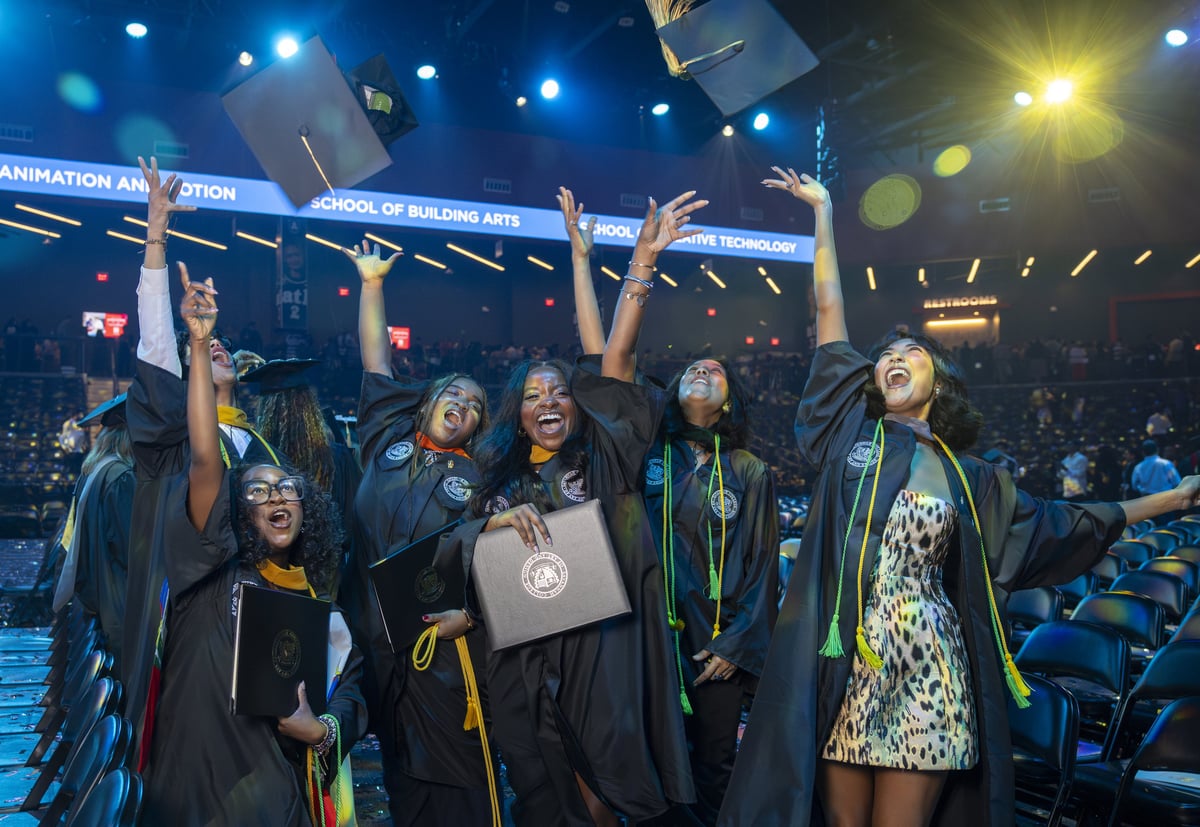 A group of graduates cheer and toss their caps in the air, diplomas in hand, under blue and amber light with the 'School of' LED ribbon wrapping the arena behind them.