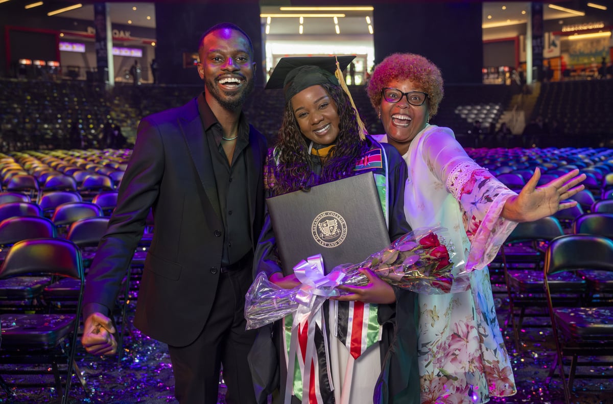 A graduate holding her diploma and a bouquet beams between two guests on the confetti-strewn arena floor after the ceremony, the emptying seats washed in purple light behind them.