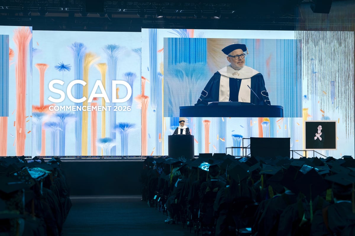 A robed speaker addresses the ceremony from the podium, projected on the large IMAG screen beside the pastel 'SCAD Commencement 2026' wall, rows of seated graduates silhouetted in the foreground.