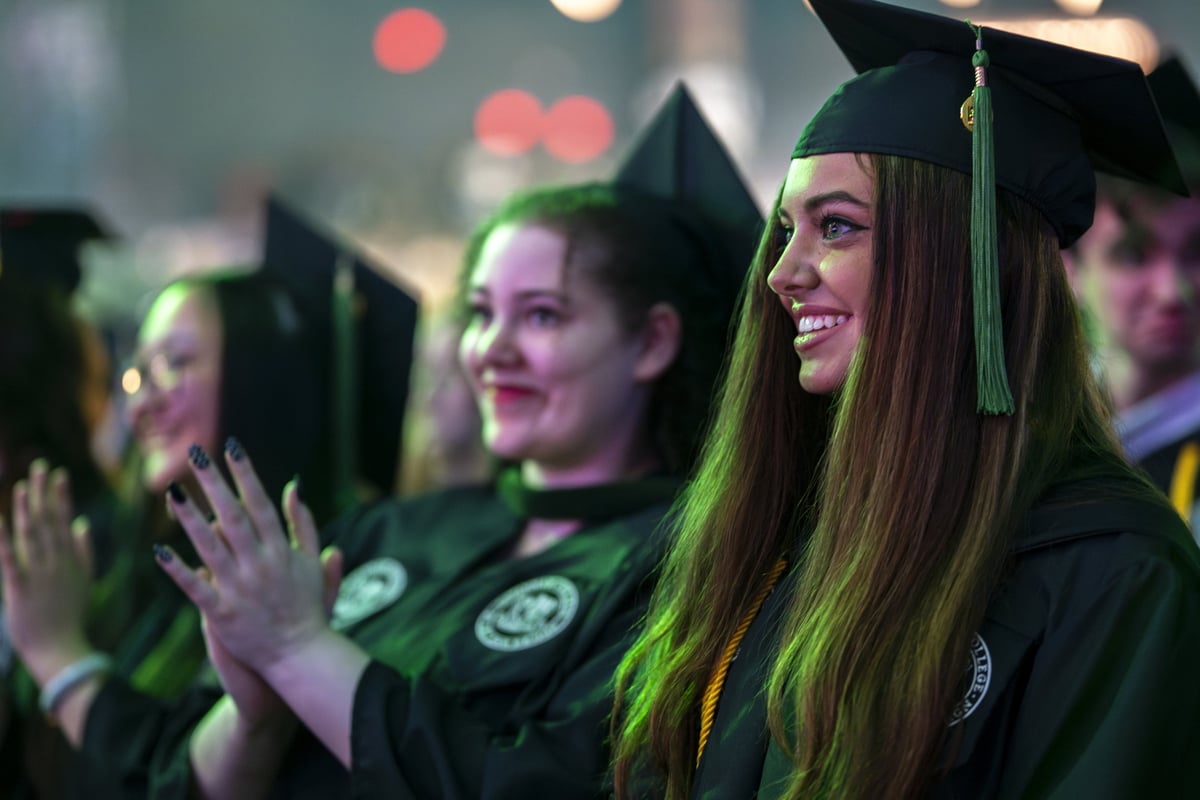 A close-up of graduates in caps and gowns smiling and applauding in the audience, lit by a soft green wash with warm bokeh behind them.