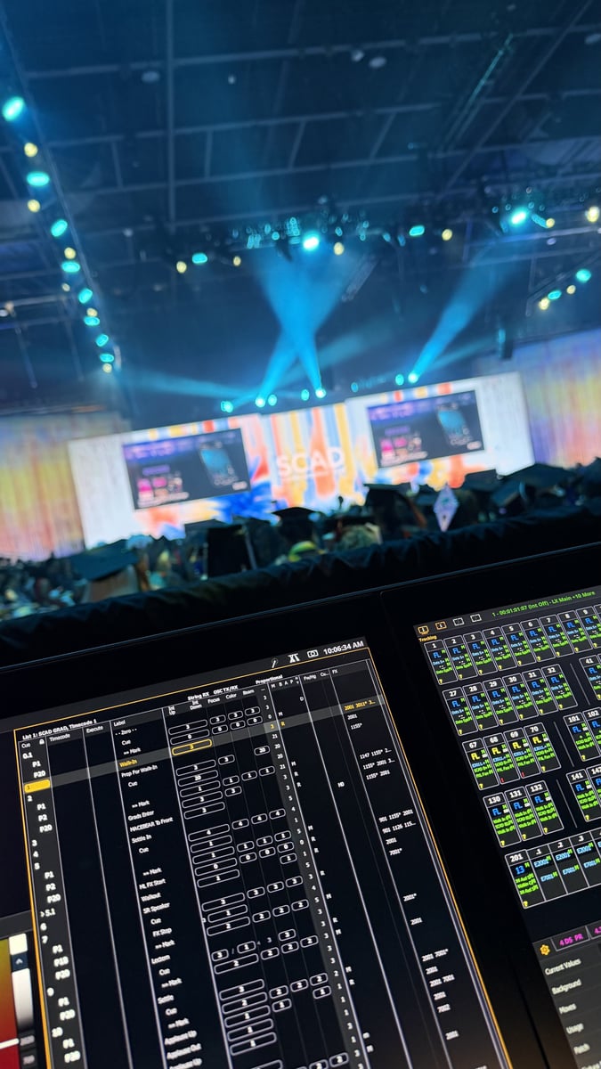 The designer's view from the lighting console during the ceremony: an ETC Eos cue list and magic sheet on screen, with the blue-lit stage, 'SCAD' screens, and seated graduates beyond.