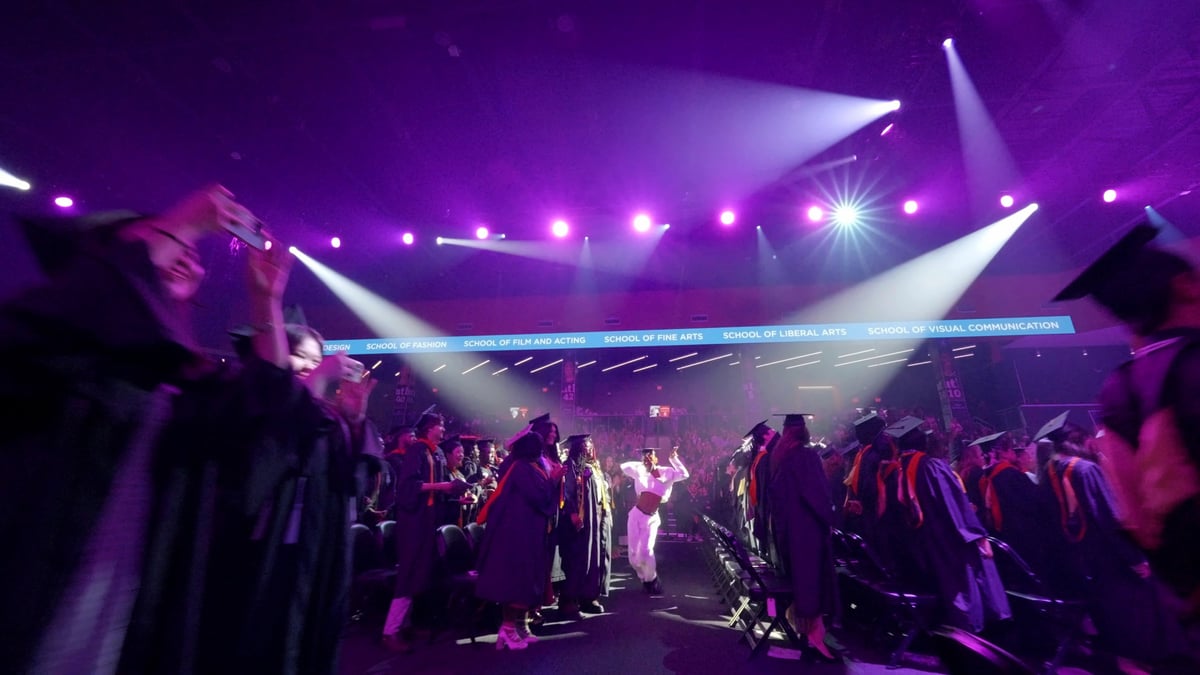 Dancers process down the arena aisle between rows of graduates under sweeping magenta beams during the commencement flashmob.