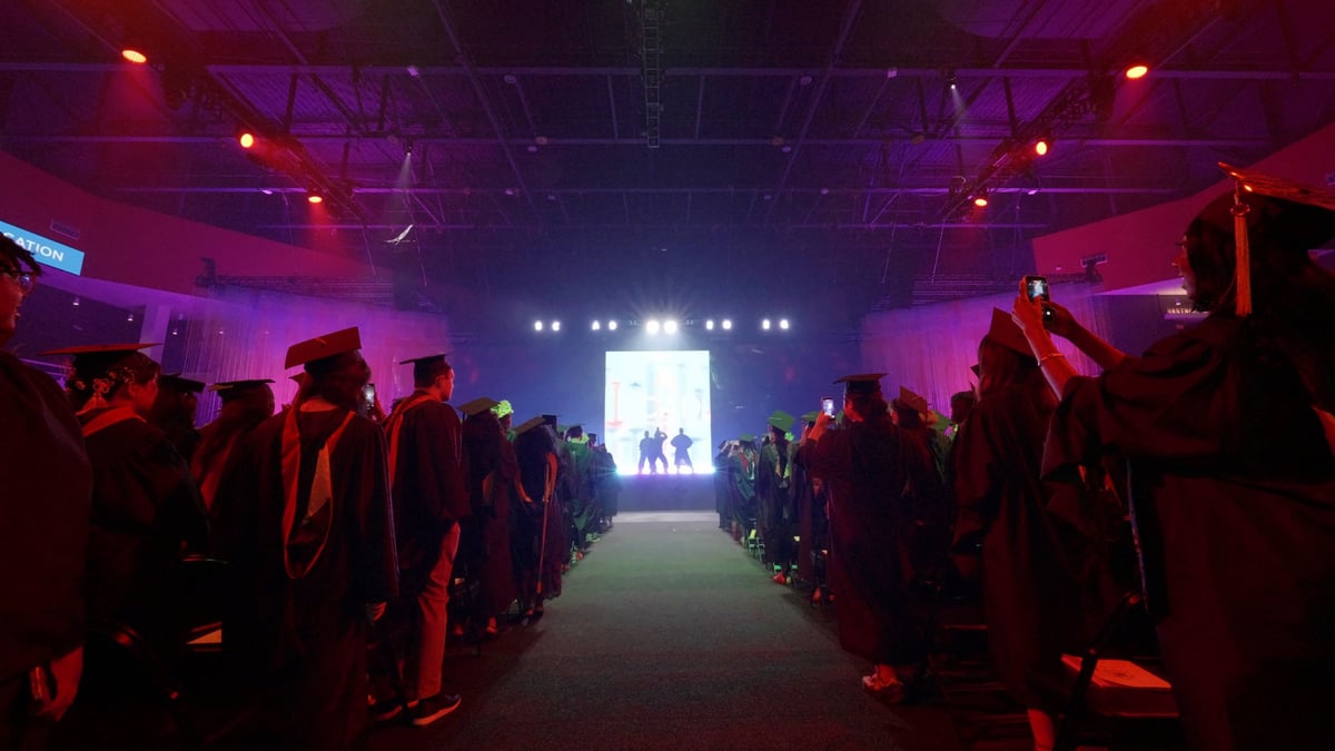 The darkened arena looks toward the distant lit stage where the flashmob dancers perform, graduates silhouetted in the foreground raising phones.