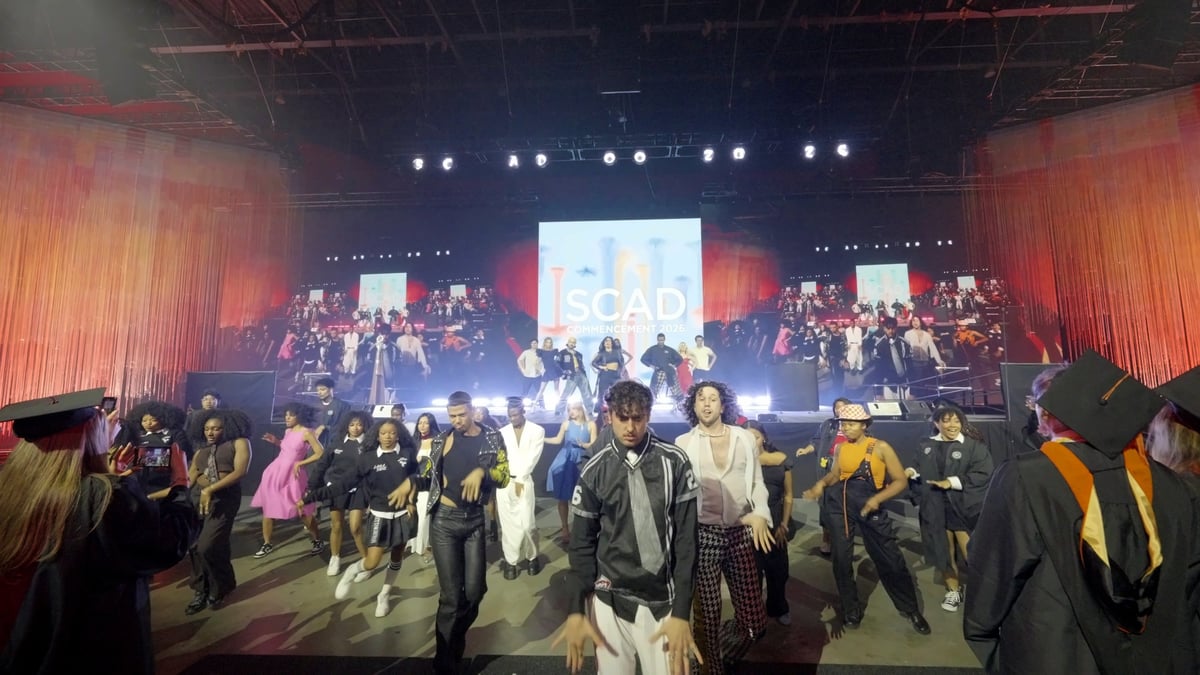 The full flashmob dance crew performs in street clothes on the main stage in front of a coral backdrop and the 'SCAD Commencement 2026' screen.