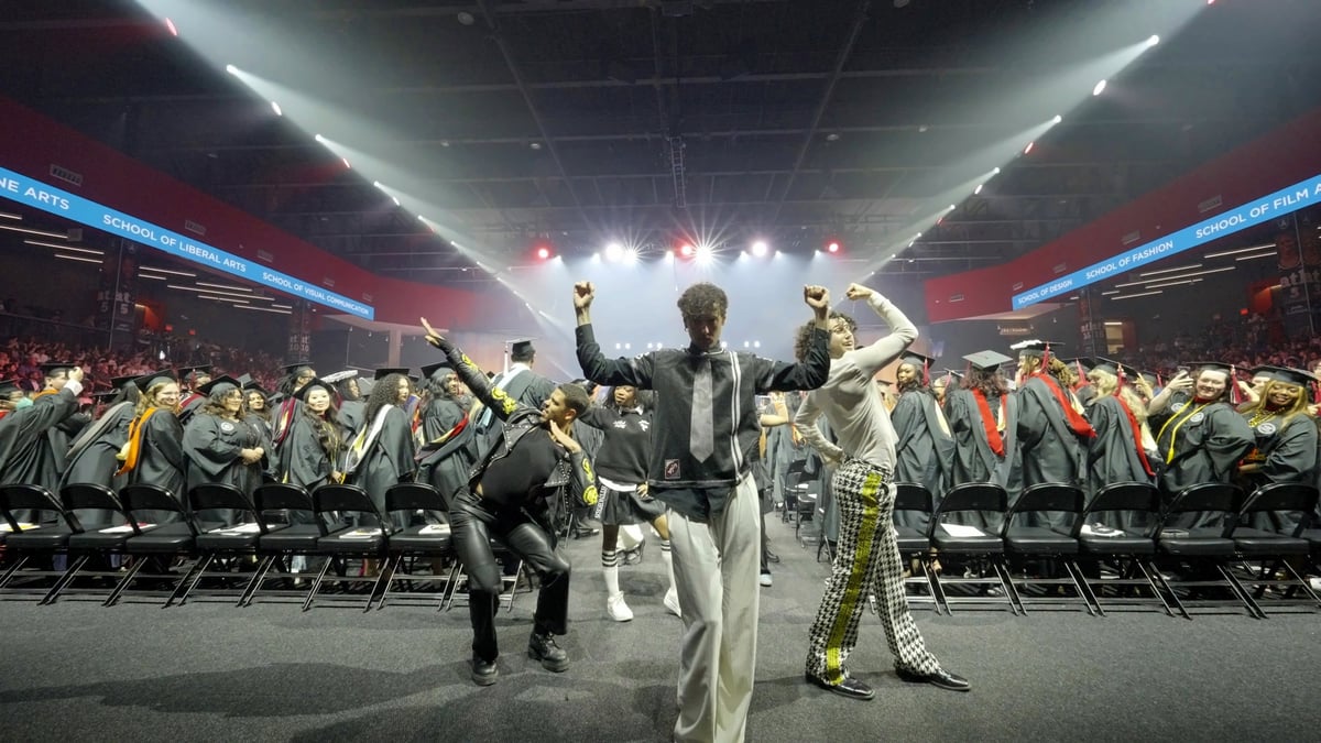 Dancers throw their arms up between the seating rows under bright white beams and red 'School of' LED ribbons as graduates cheer around them.