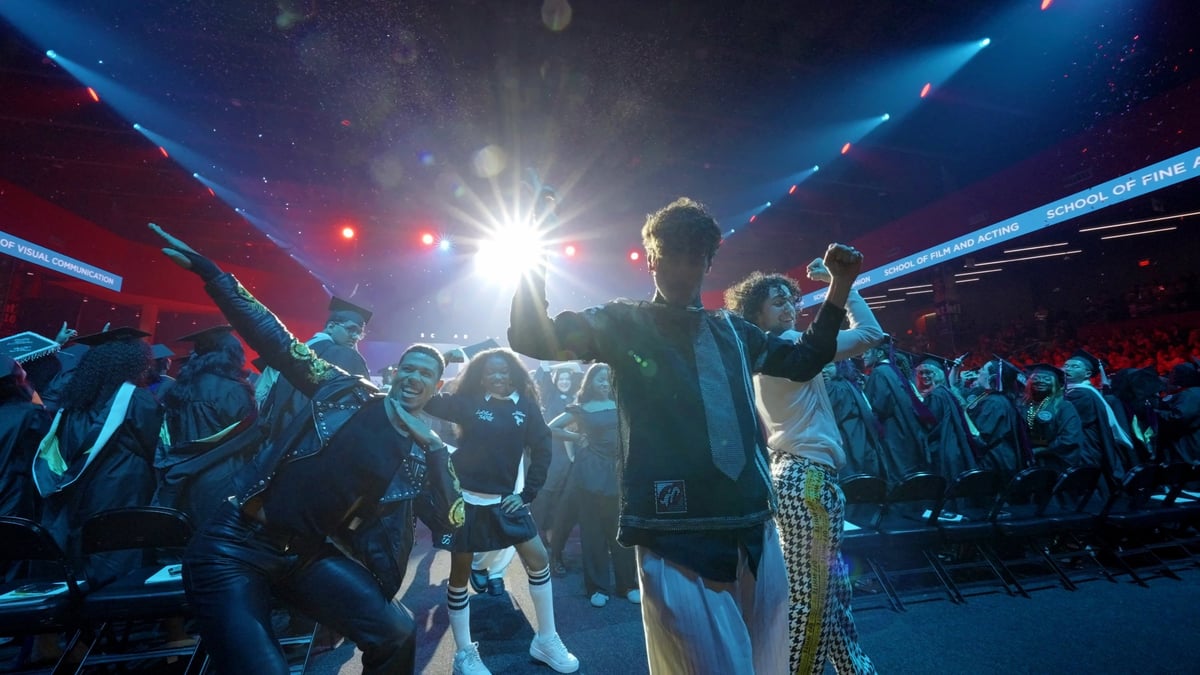 A dancer raises a fist beside a burst of white light, blue and red beams crossing the arena as graduates dance in the aisle.