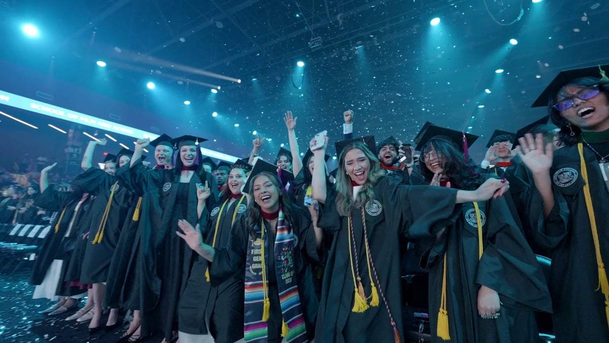 Graduates in caps and gowns cheer toward the camera under a teal wash as confetti falls at the end of the flashmob.