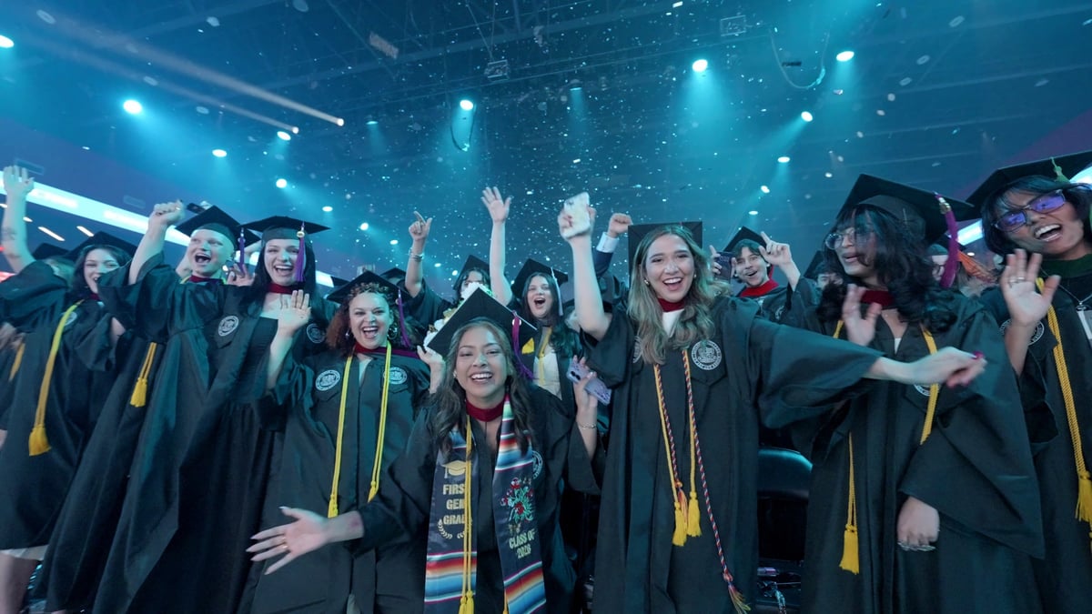 A packed row of graduates throws their hands up, cheering under teal light and drifting confetti.