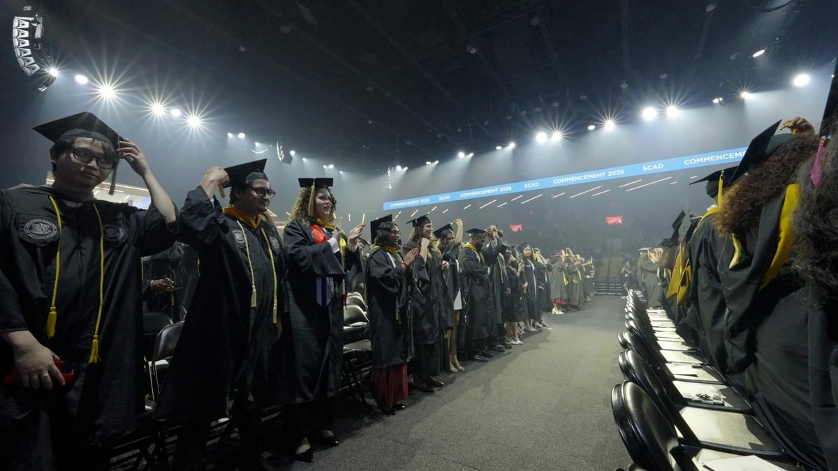 Graduates rise from their rows as the flashmob kicks off, white light and haze filling the arena beneath the 'SCAD Commencement 2026' ribbon.