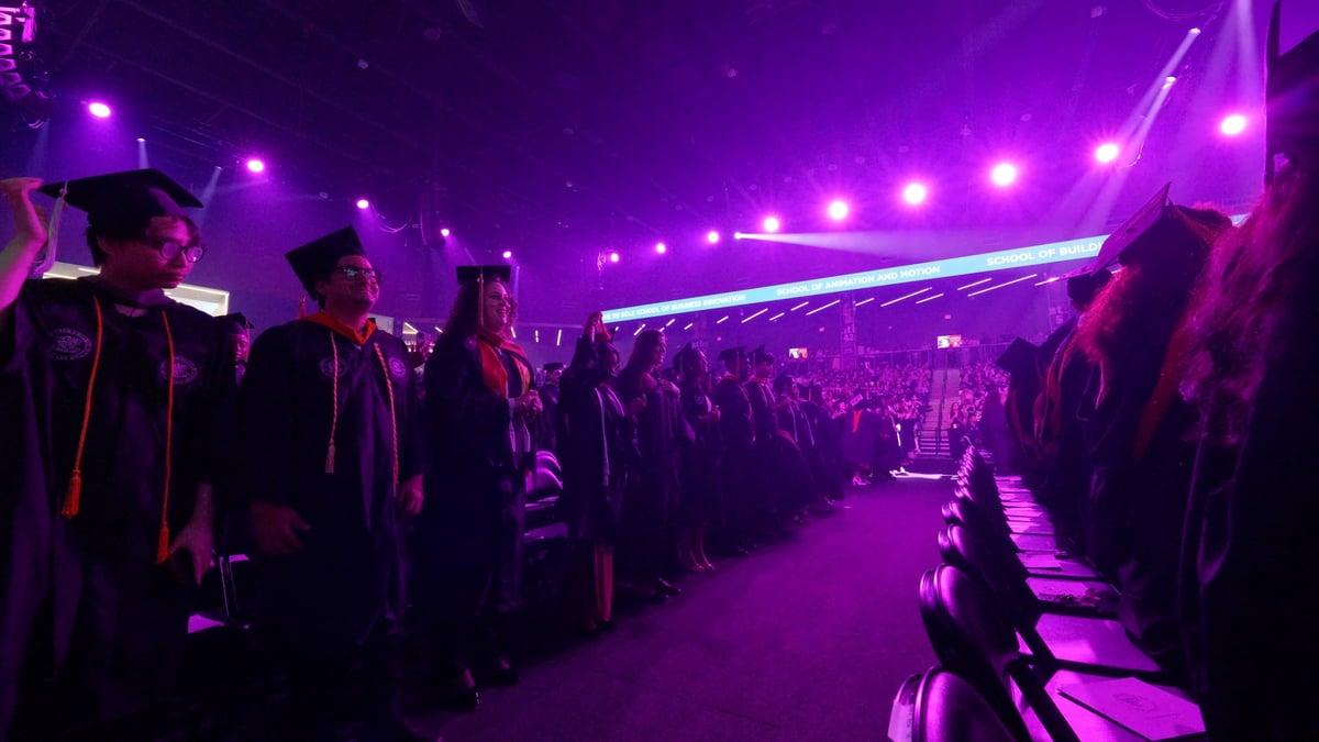 Rows of graduates stand watching the flashmob under a saturated purple wash, the LED ribbon and packed arena bowl glowing behind them.