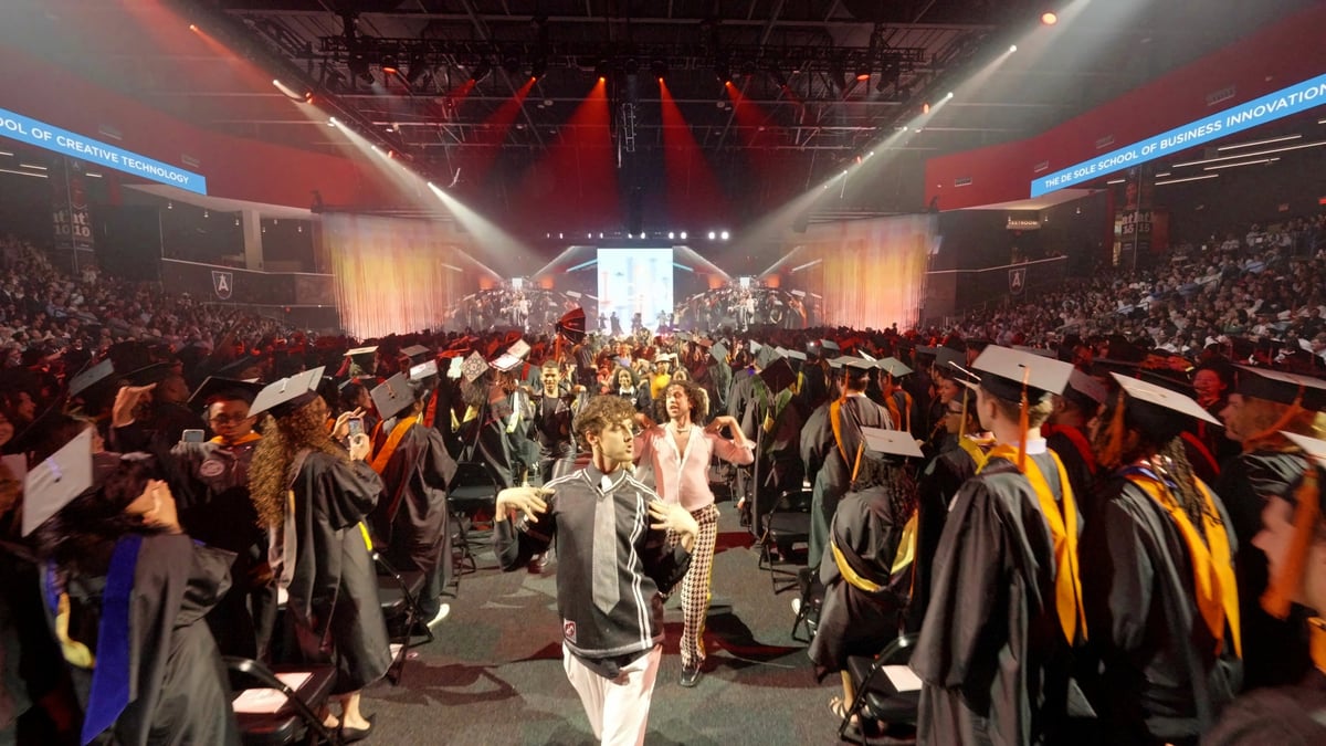 A flashmob dancer performs in the center aisle among the seated graduates, red wash and white beams sweeping the arena toward the distant stage.