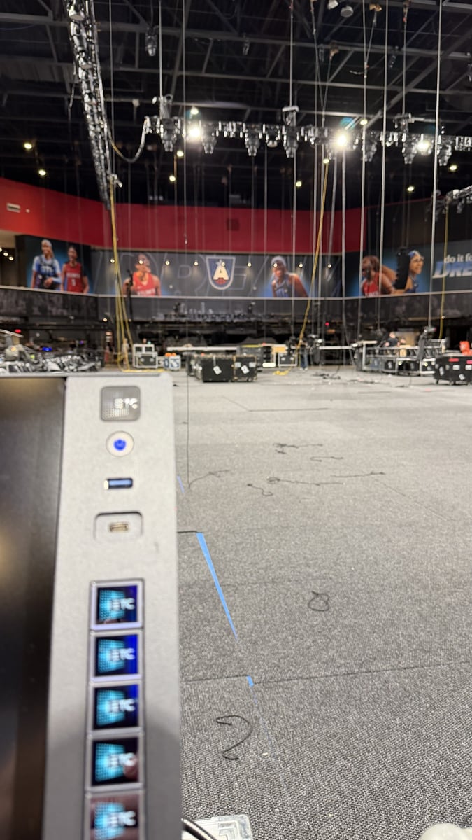 Load-in at Gateway Center Arena: an ETC lighting console in the foreground with rigging motors, road cases, and the empty arena floor marked with spike tape behind it.