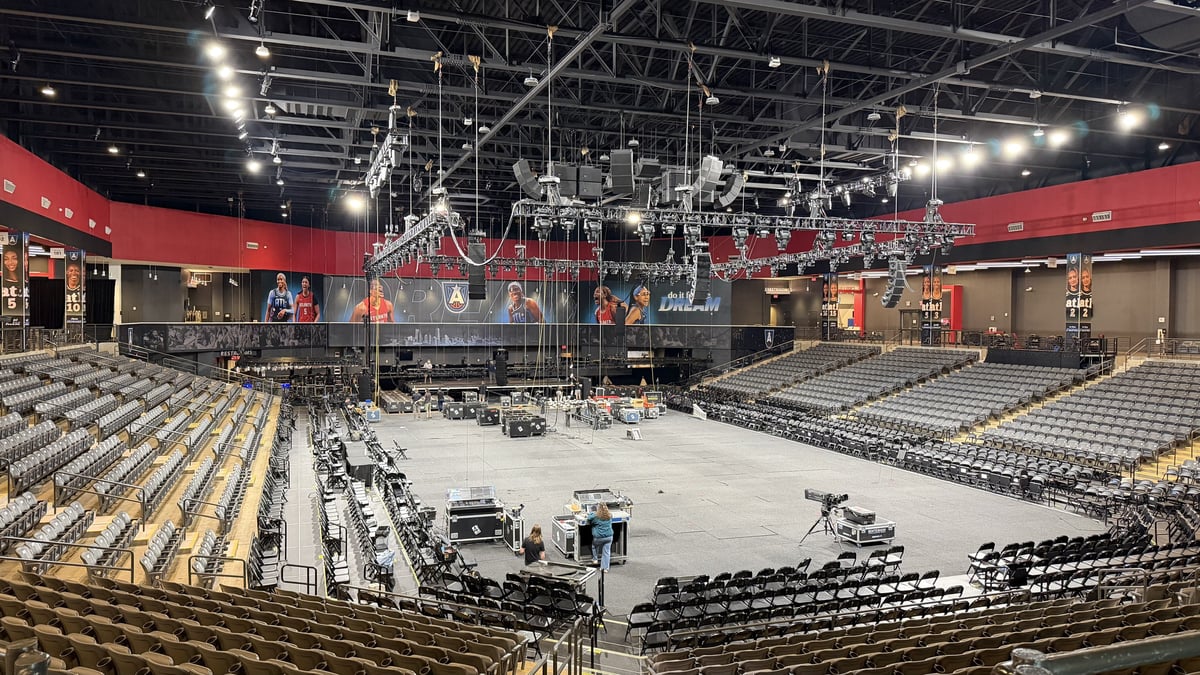 A wide view of Gateway Center Arena mid load-in, the central stage truss half-built, floor chairs going in, road cases and a camera position on the arena floor under the lighting grid.