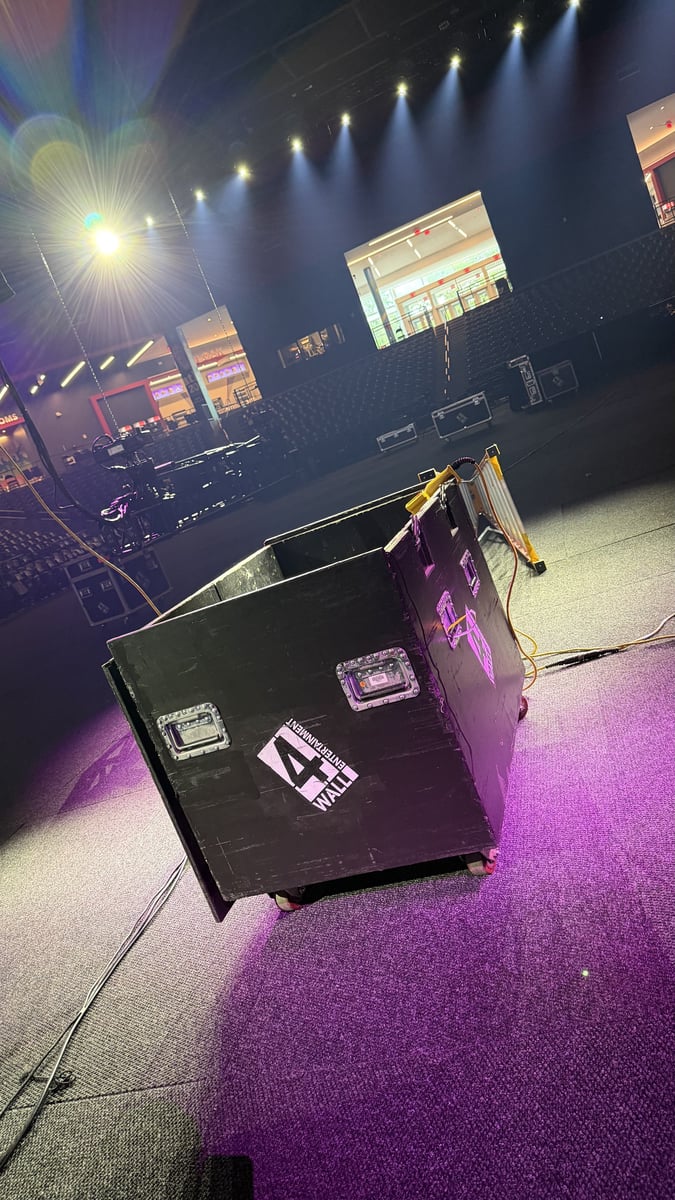 A 4Wall road case on the arena floor as the rig warms up in a magenta wash, cable runs snaking across the carpet and the empty seating bowl beyond.