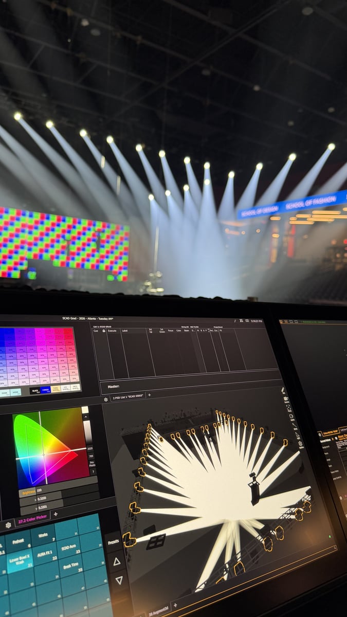 The view from the programming position: an ETC Eos magic sheet and color picker on the console screens while the rig fans white beams through haze over an RGB-test LED wall during tech.