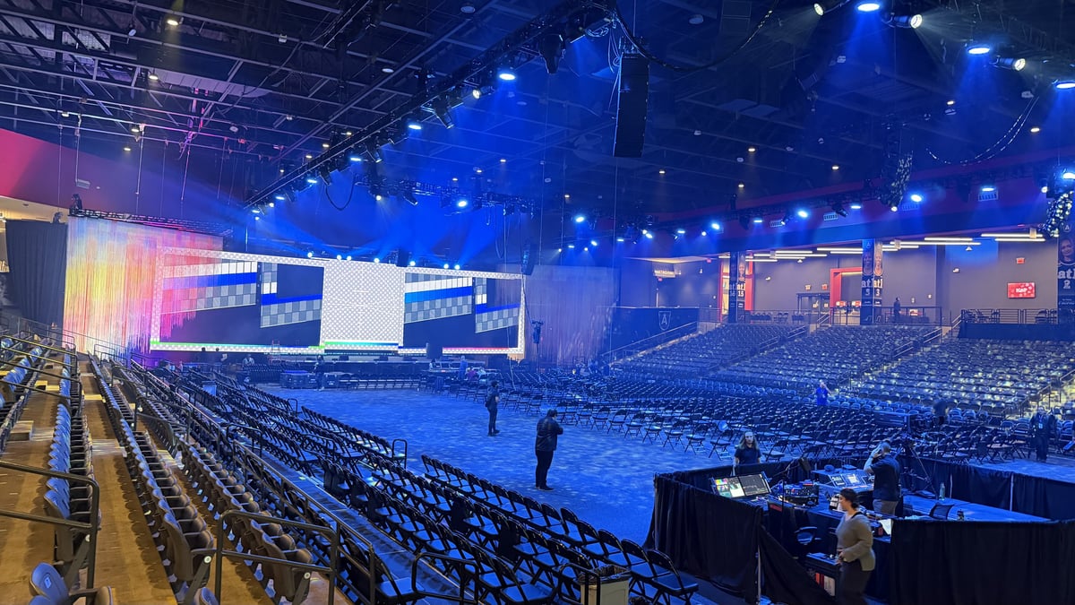 The arena set for the ceremony under a deep blue wash, rows of chairs in place, LED screens running a test pattern, and crew at the front-of-house tables before doors.