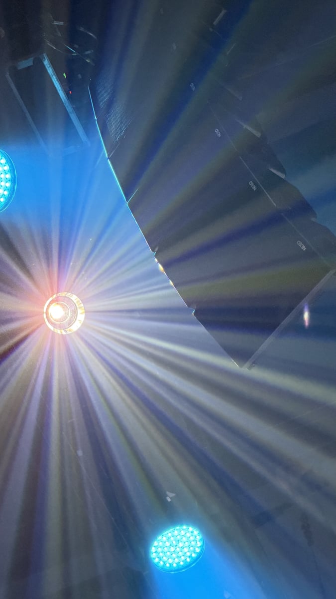Looking straight up into the rig as moving lights burst through the haze, a sunburst flare from one fixture and blue beams raking past a flown line-array speaker.