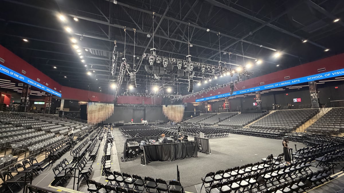 The finished setup under work lights: the central lighting rig hung over the stage, chairs set across the arena floor, the front-of-house table skirted, and the 'School of' LED ribbon wrapping the room.