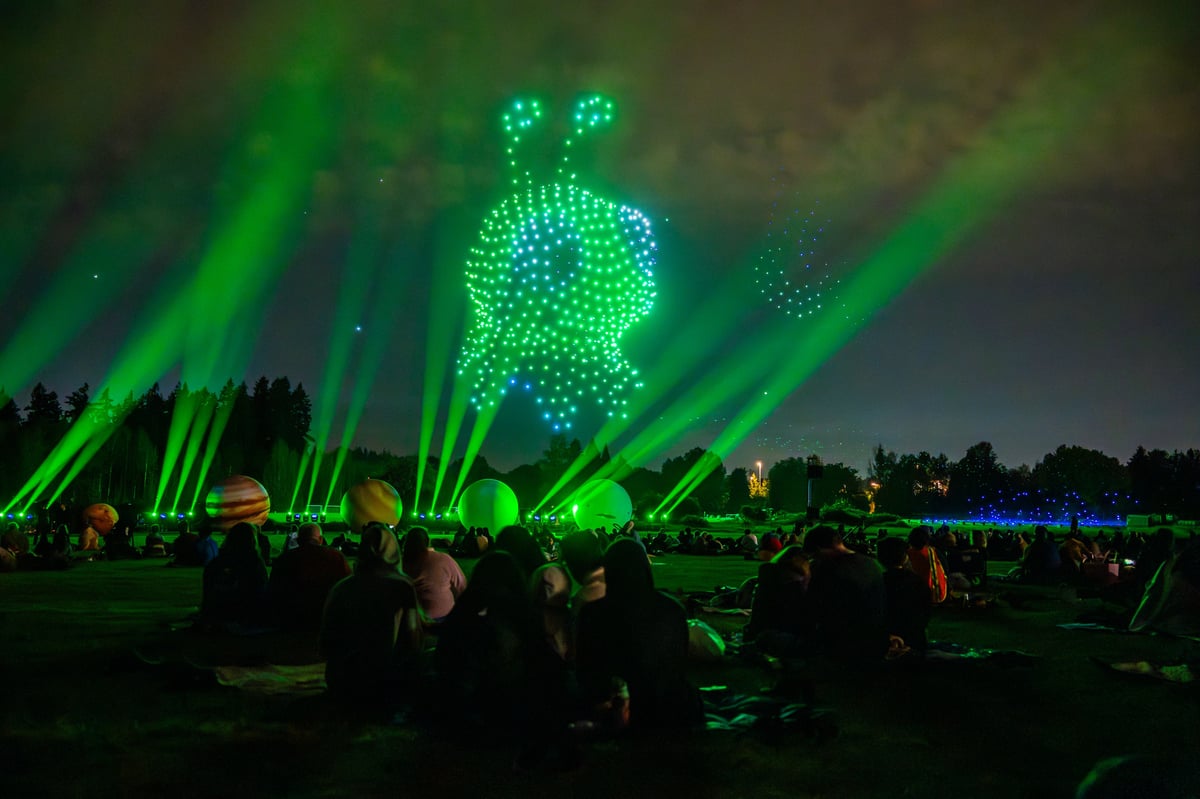 Freddie, the show's alien hero, rendered in green drones with two antennae above the crowd as green beams fan up past glowing planet spheres.