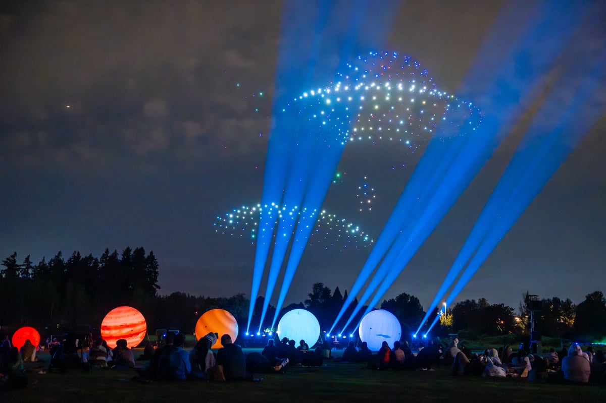 A spaceship rendered in blue and white drones hovers over the field, blue beams fanning up from the stage past a row of illuminated planet spheres.