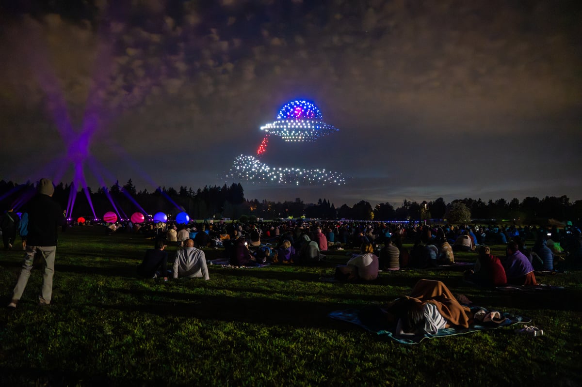 A flying saucer drawn in blue drones with a red tractor beam over the crowd, purple beams sweeping the sky above glowing planet orbs.