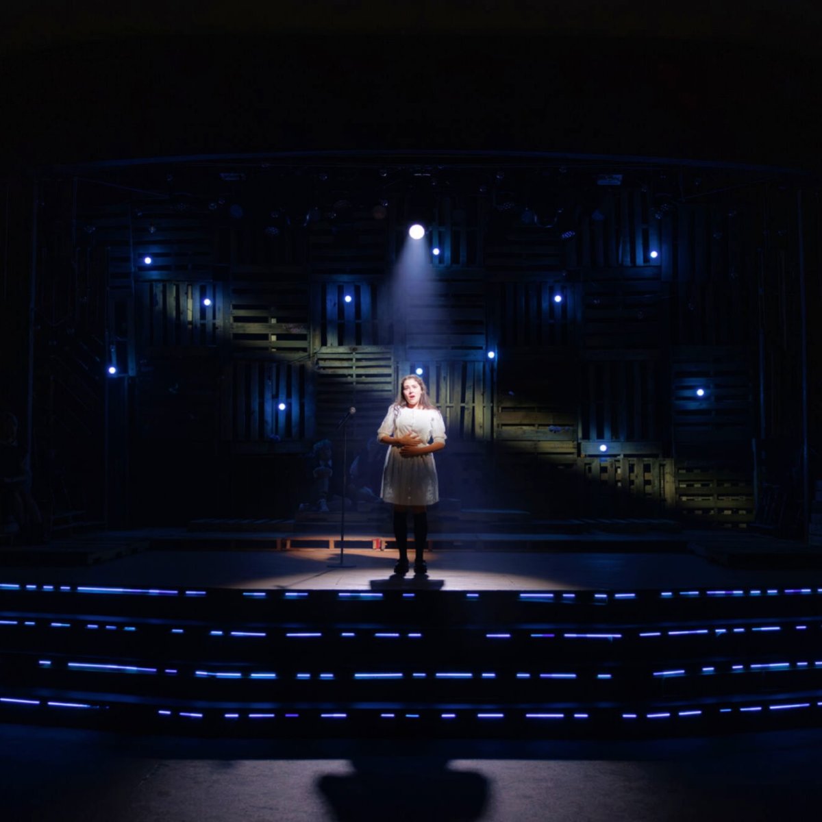 A young woman in a white dress sings alone at a microphone in a tight spotlight, the pallet-wood wall behind dotted with blue pinlights and blue LED striping along the steps.