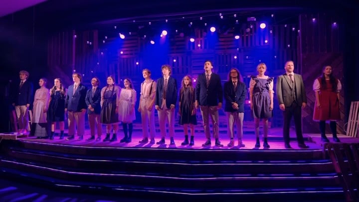 The full company stands in a line across the stage in a purple and magenta wash, the pallet-wood wall rising behind them.