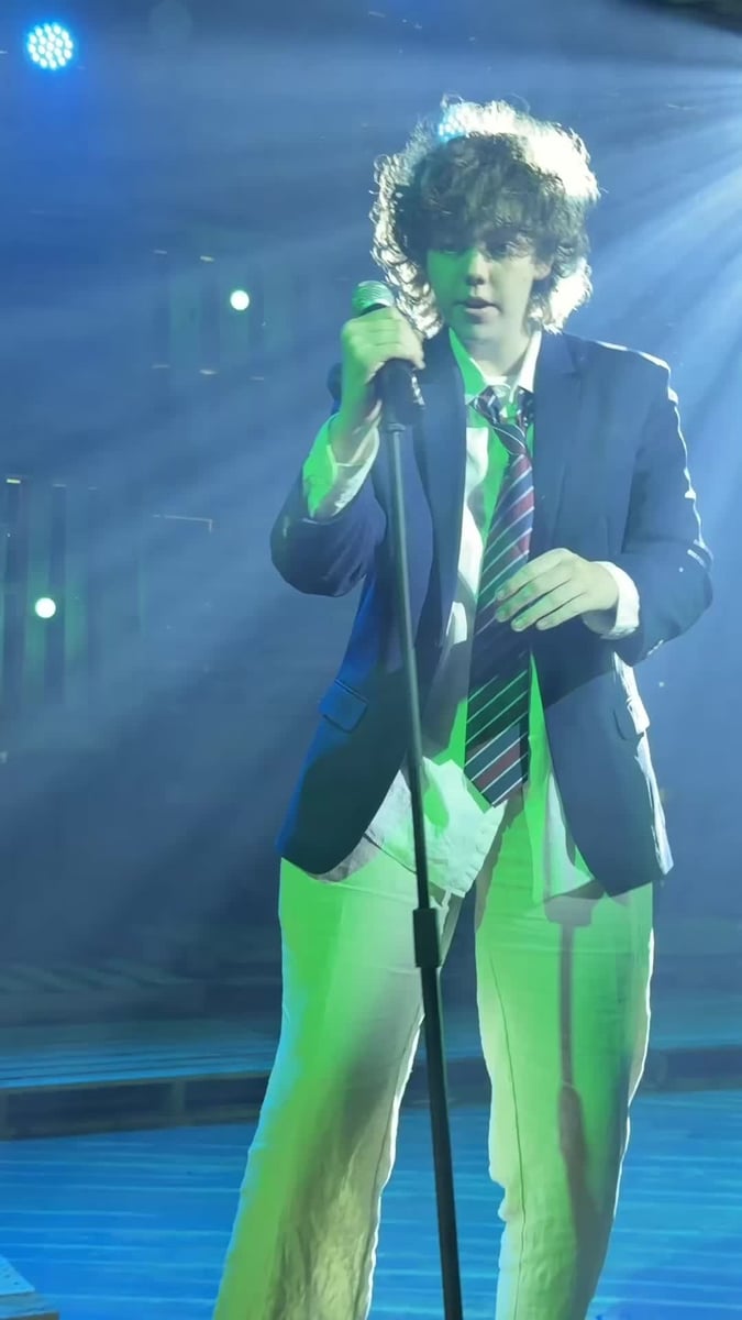 A young man in a school blazer and striped tie sings into a handheld microphone, lit in green and blue amid haze.