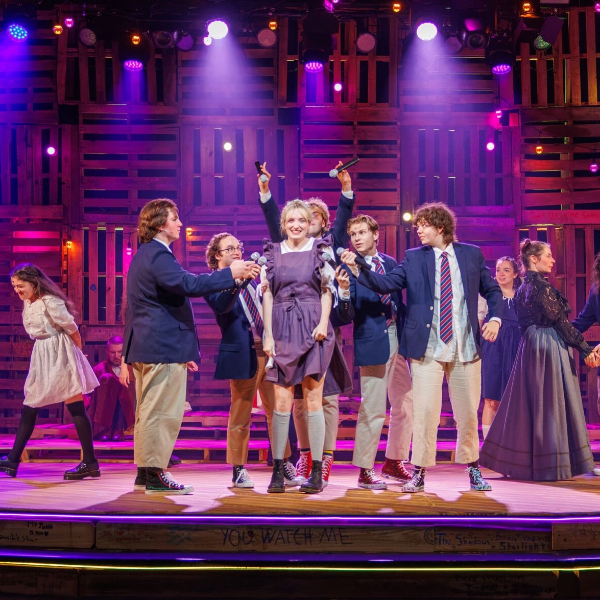The company clusters around a young woman in a denim dress at center, microphones raised overhead, in a magenta wash on the graffiti-marked pallet stage.