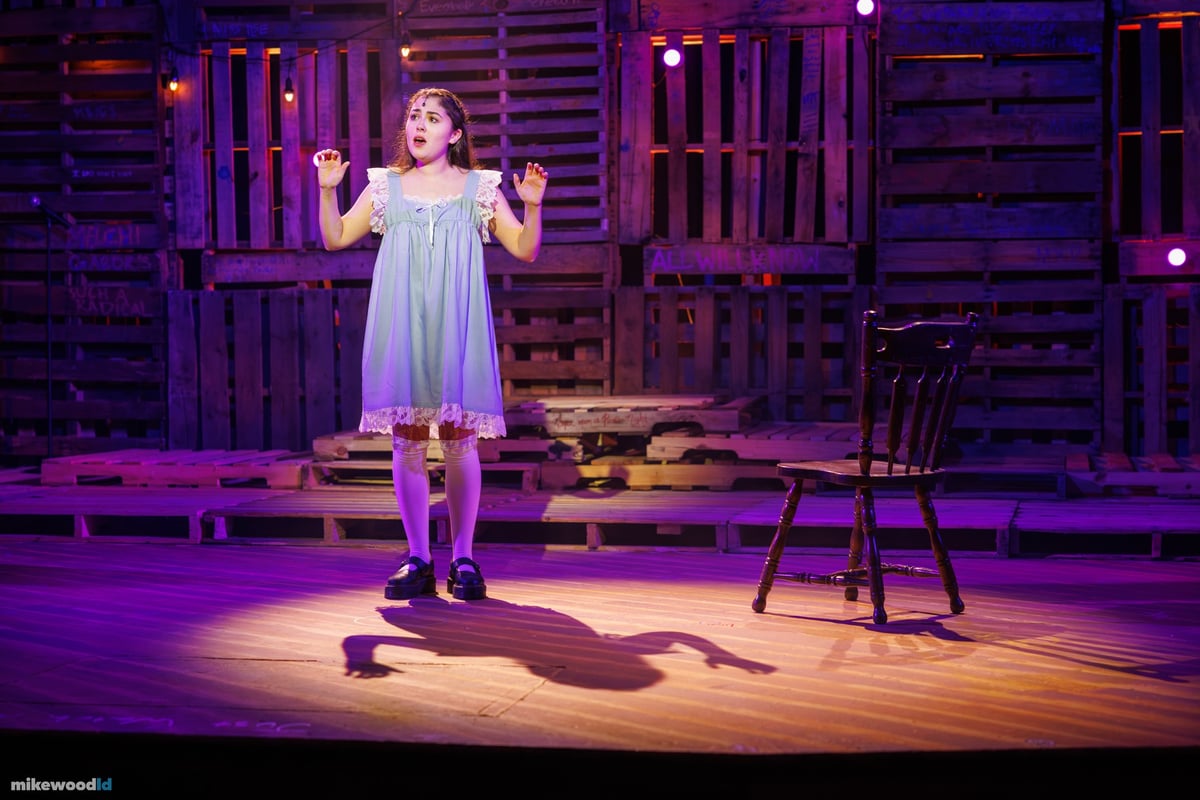 A young woman in a pale blue slip dress stands alone in a lavender wash, arms raised, her shadow thrown long across the plank stage.