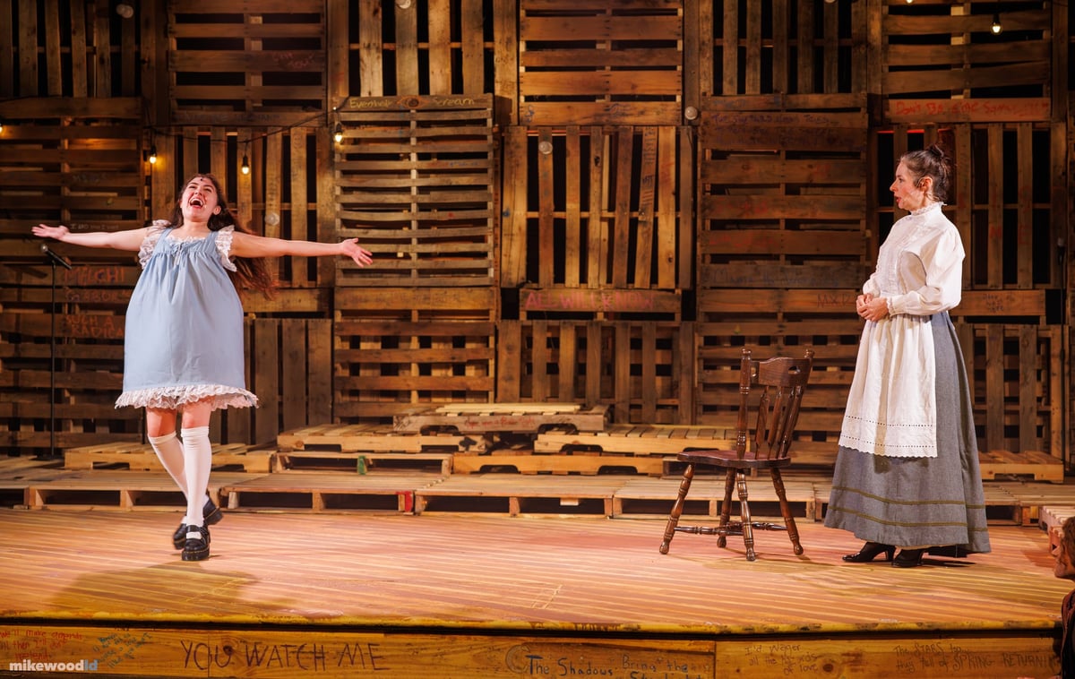 A young woman in a blue slip dress skips across the stage in warm light as a woman in a period dress watches from the pallet wall.