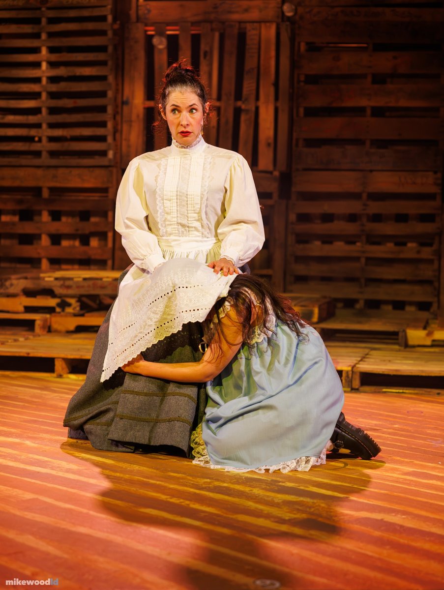 In warm amber light, a woman in a white high-collar blouse kneels and cradles another woman lying on the stage floor.