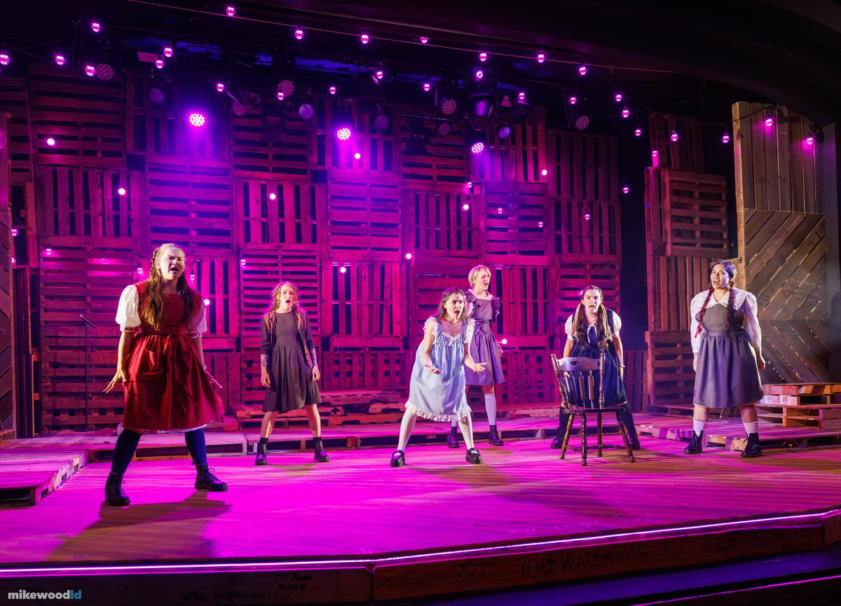 The young women of the company line the stage in period school dresses under a saturated magenta wash.