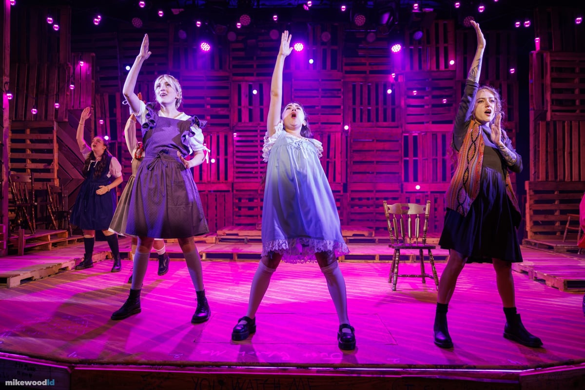 Three young women throw their arms overhead in a hot pink wash, the rest of the cast behind them on the pallet-wood set.